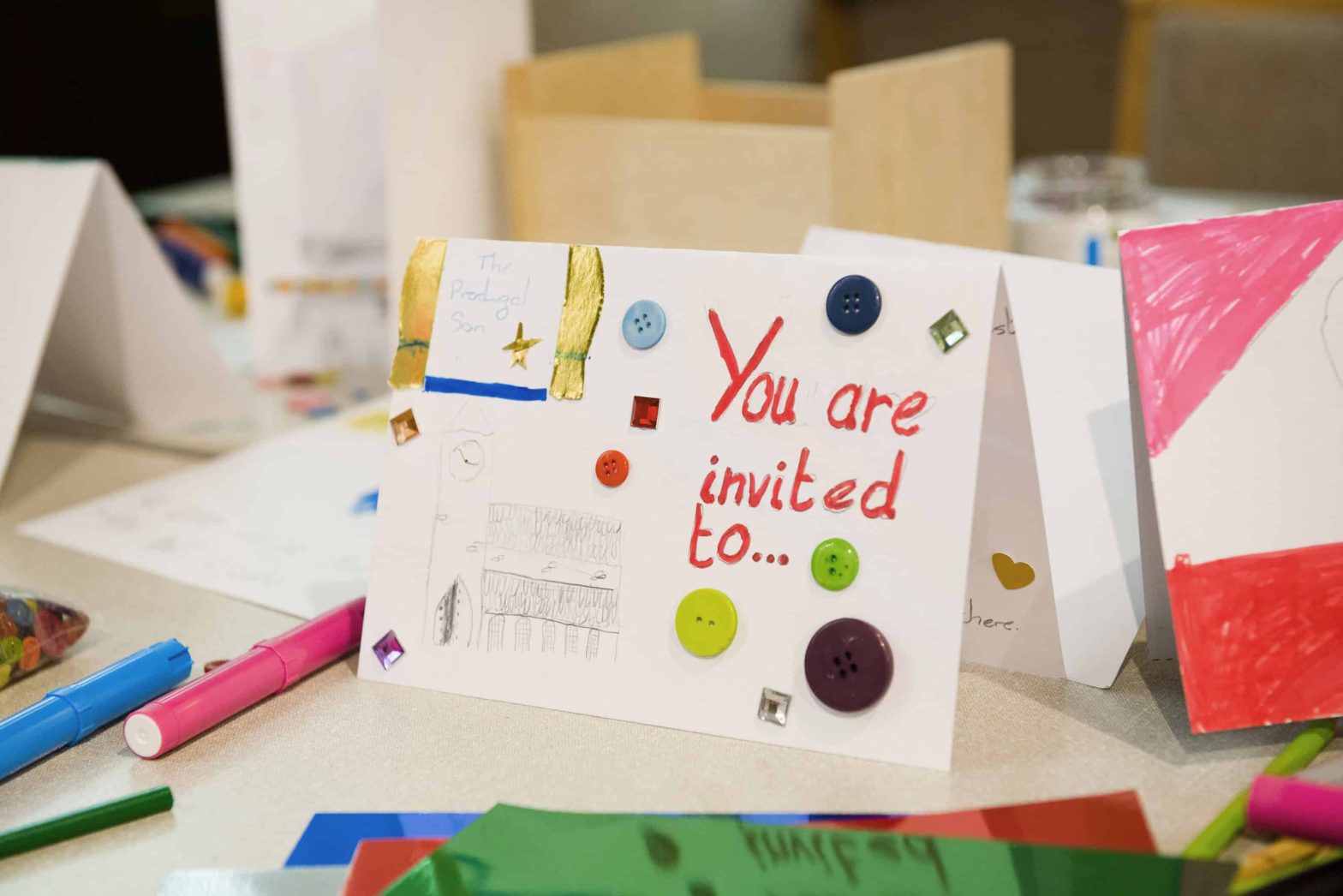 A Childs home made card with 'You are invited to...' written on the front. Placed on a table with craft materials on it.