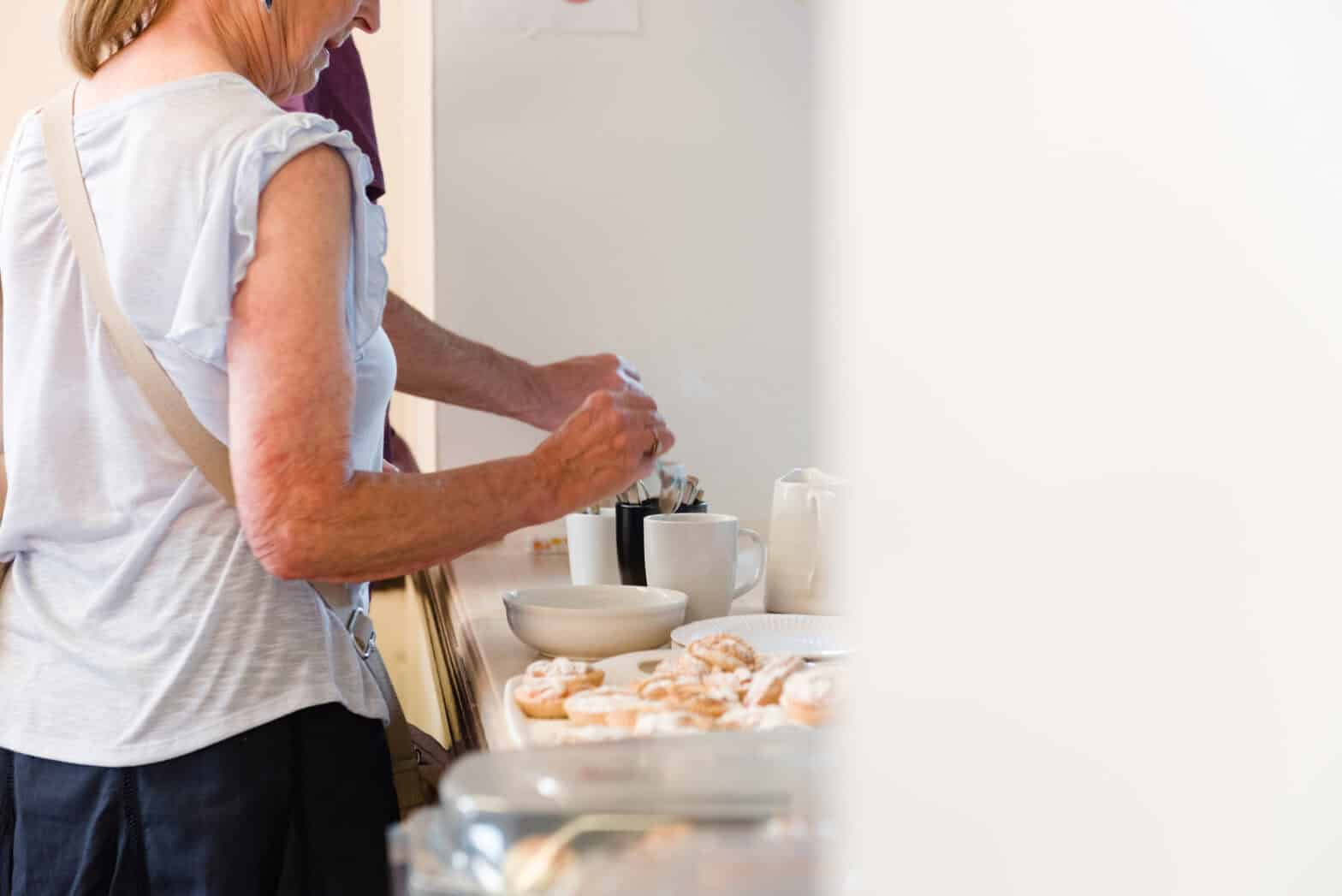 An older woman helping herself to a cup of tea and some cake from a self-service table.