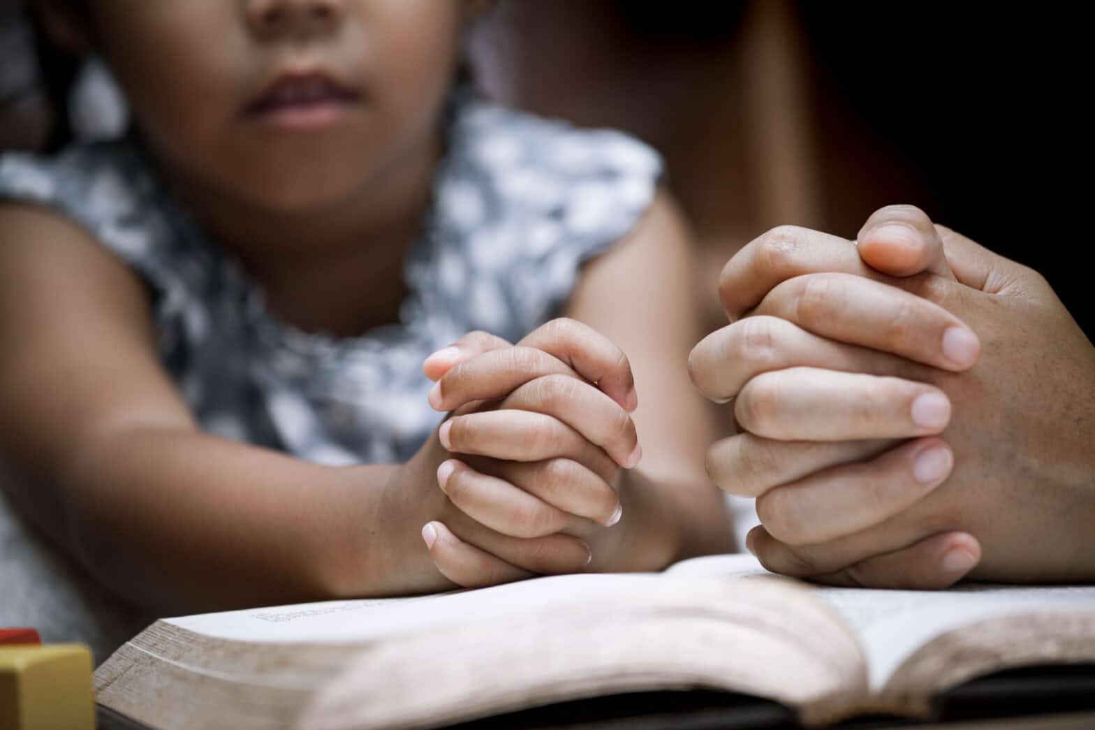 Mother and little girl hands folded in prayer on a Holy Bible together.