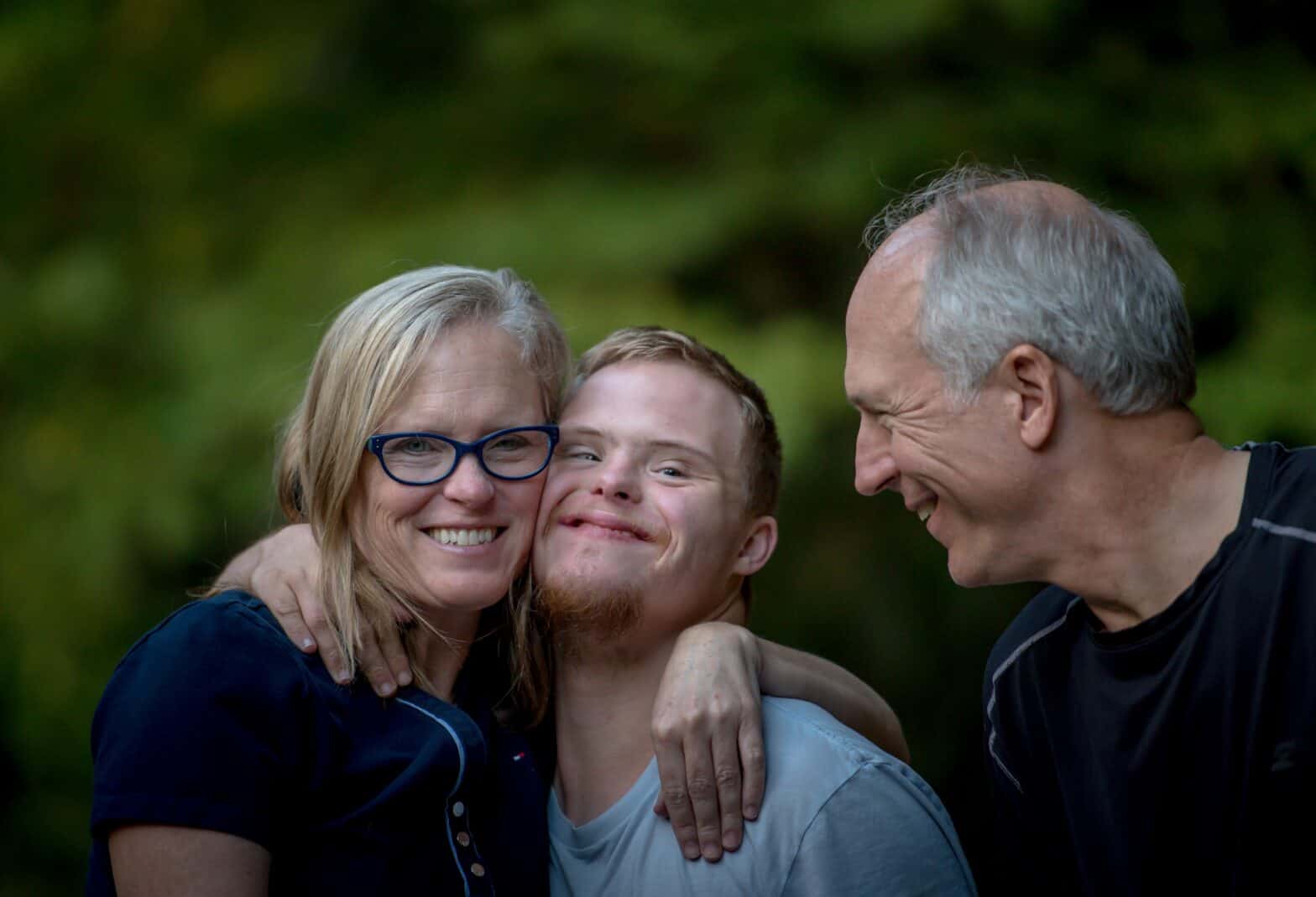 Three adults standing close together, in the centre a young adult with Down Syndrome is hugging his mother, his father the other side looks on smiling.