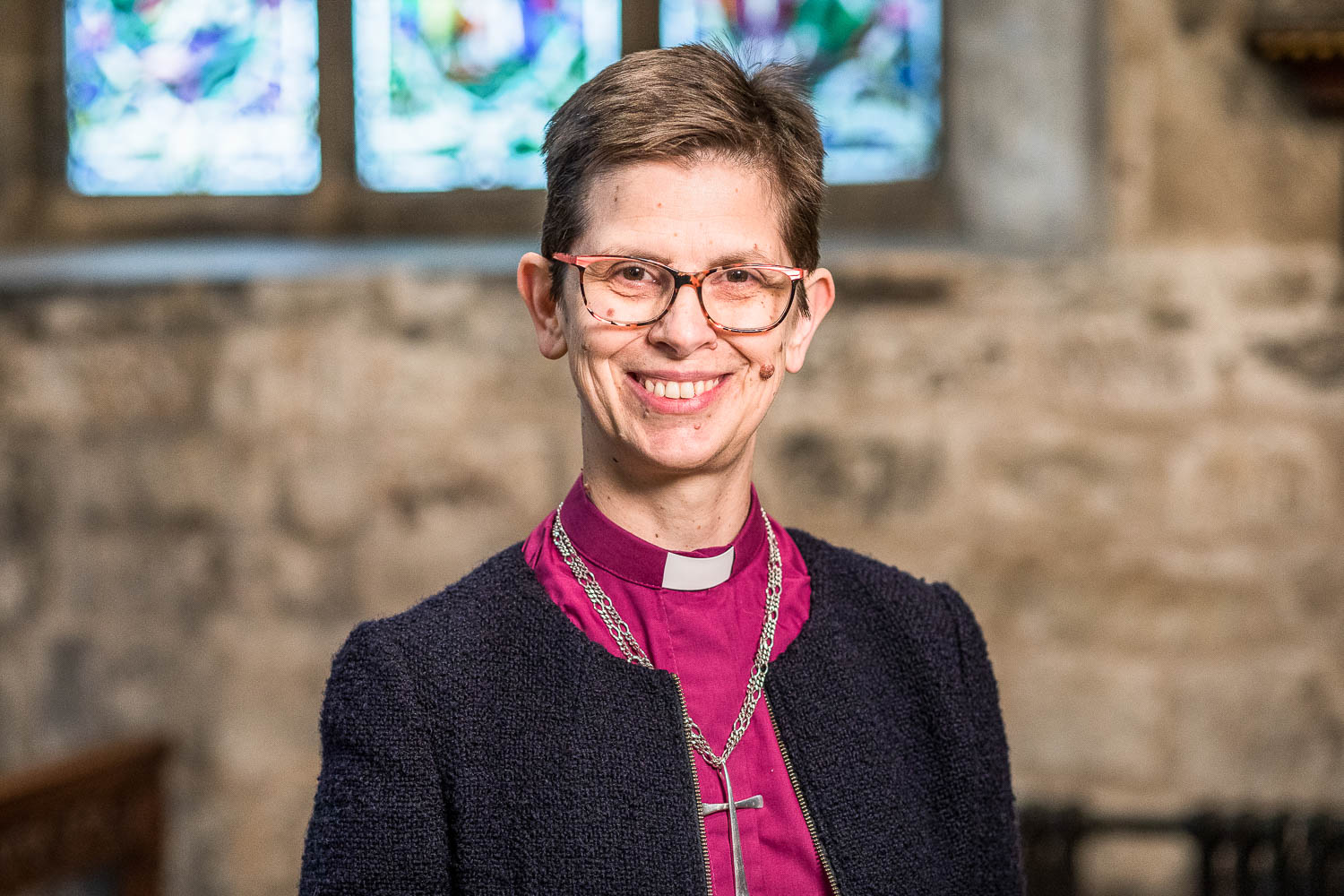 A portrait of Libby Lane, Bishop of Derby in a church. She wears a purple clerical shirt, with collar, gold cross, and suit jacket. She is smiling to camera.