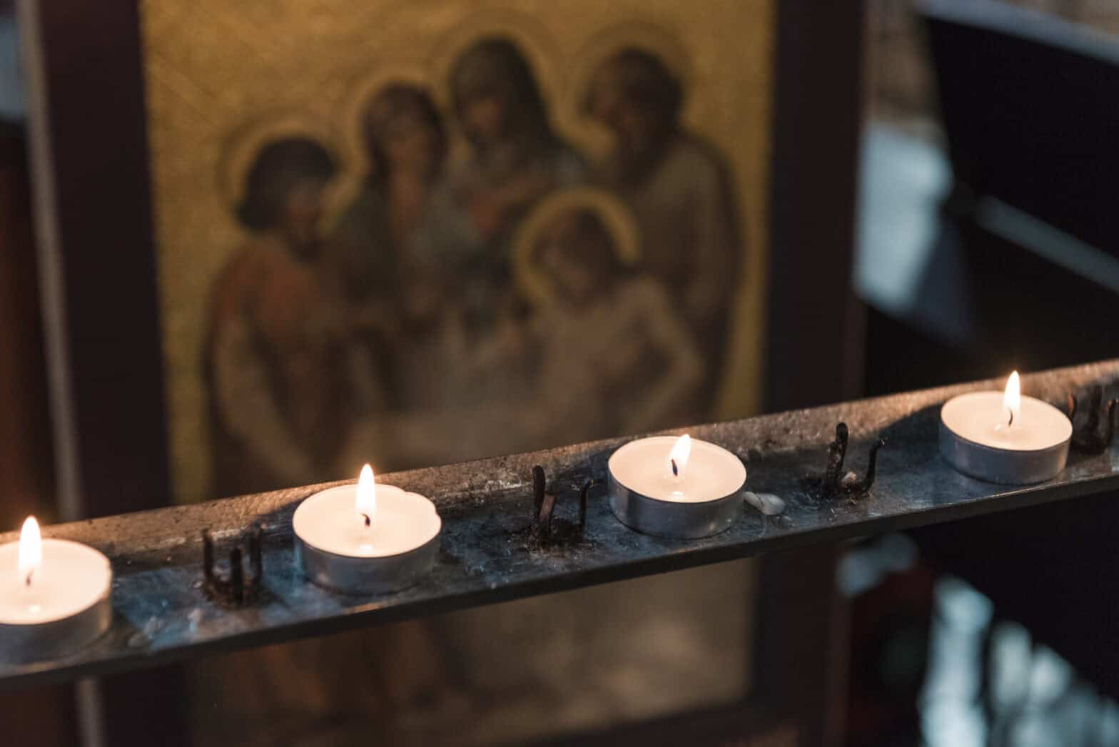 Tea-light candles lit on an iron railing in church, in the background there is a religious painting.