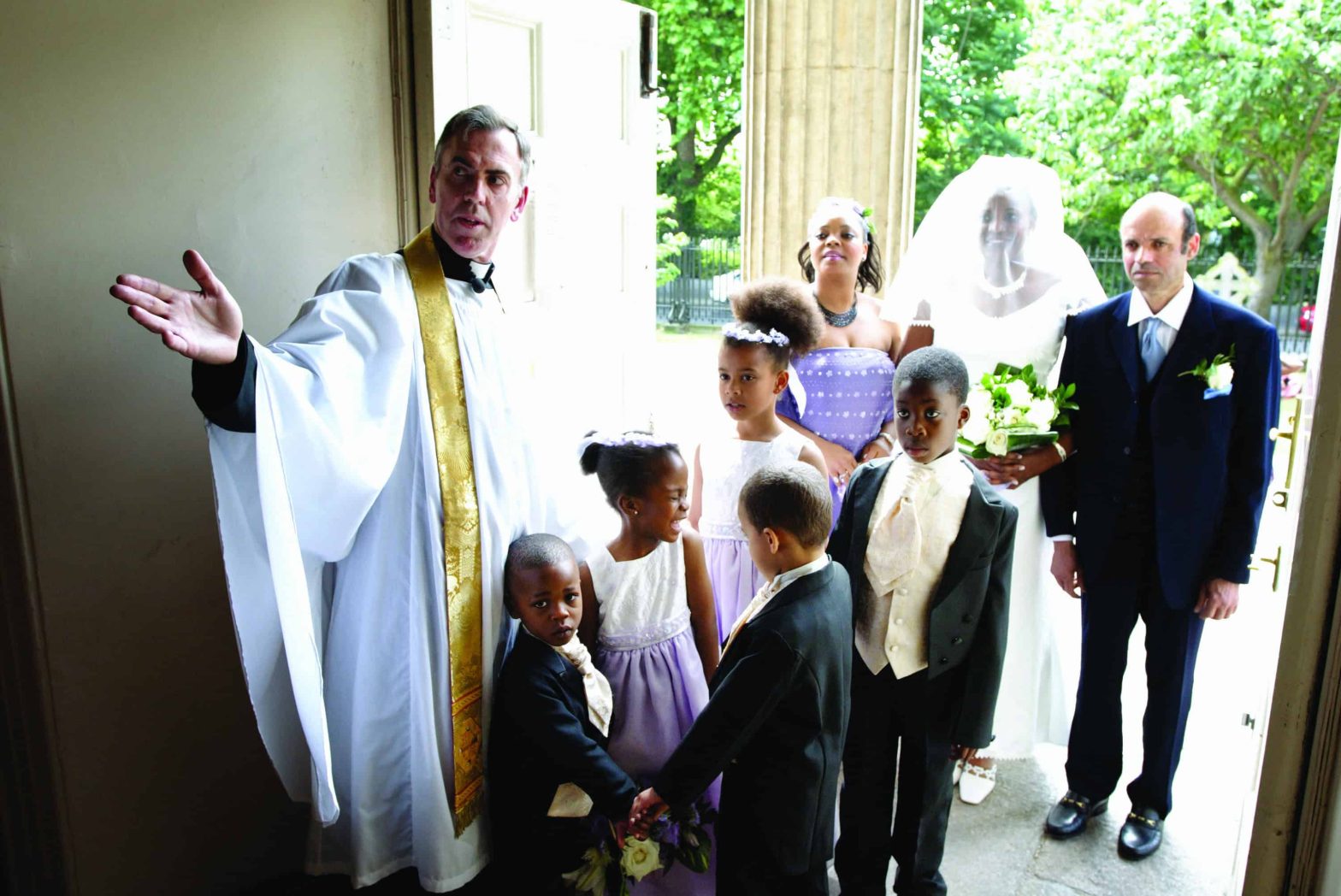 Male vicar showing a bride, her father, and young bridesmaid and page boys into church.