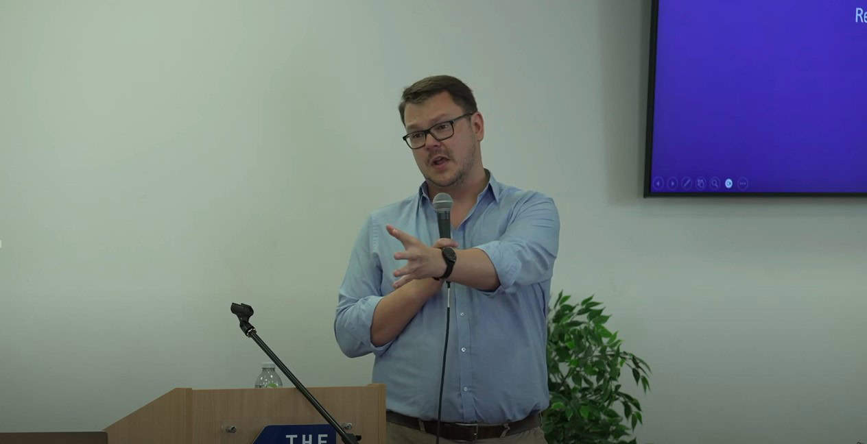 A man with short hair and glasses holder a microphone, his other hand reaches out in front of him.