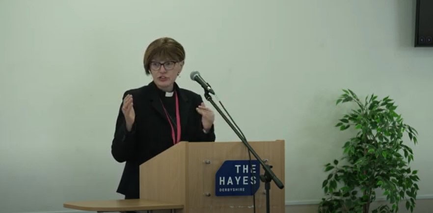 A lady wearing a clerical collar and black suit talking at a lecture. On the front is a sign that reads 'The Hayes'.