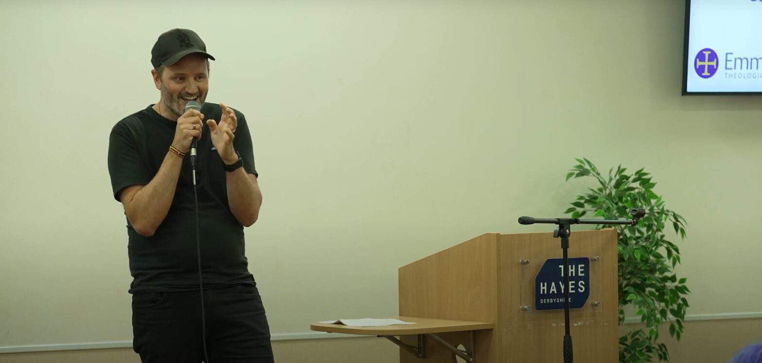 A middle aged man wearing a black baseball cap, t-shirt, and jeans, talks animatedly into a microphone, Next to him a lecture with a sign saying 'The Hayes'.