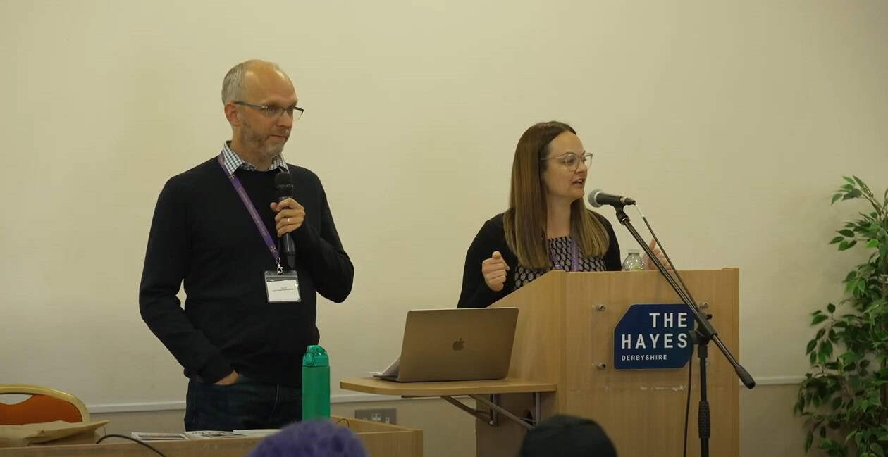 An older man holding a microphone, an a woman talking into a microphone. On the lecture is the name 'The Hayes'.