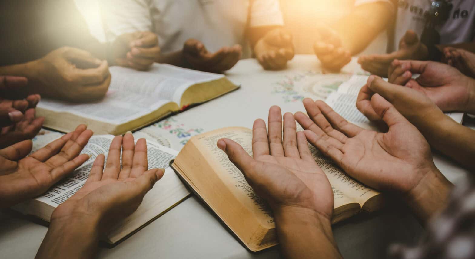 People praying around a table with their hands open on top of open bibles. The image is cropped to focus on the open hands.