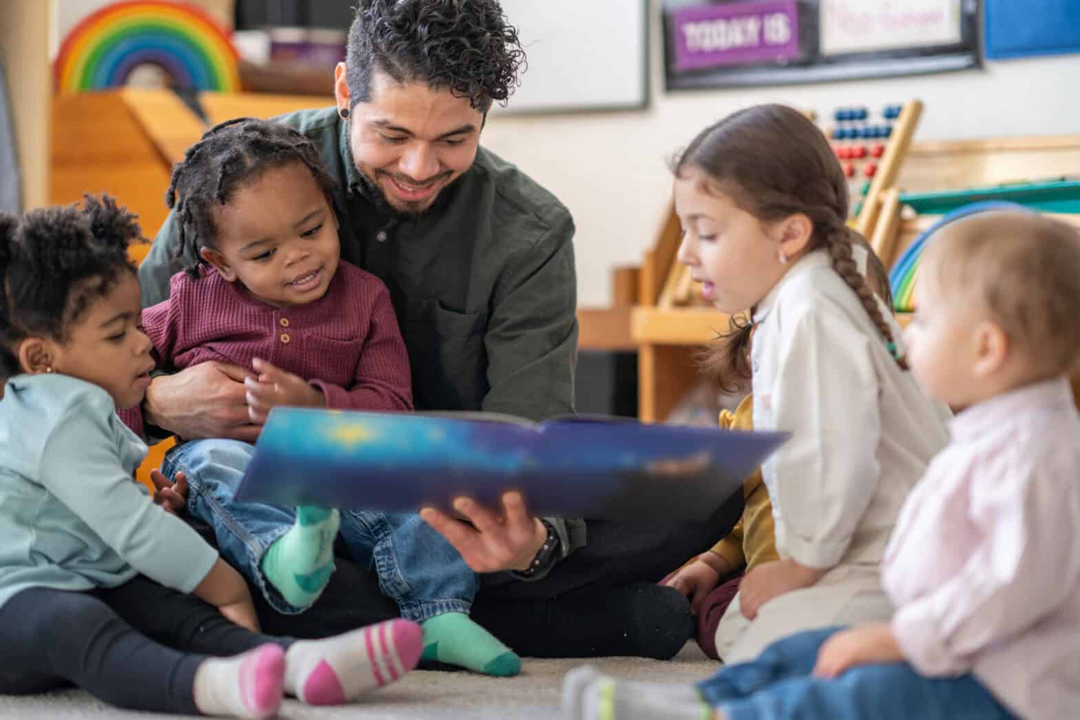 A male adult sits on the floor with a child on his lap and other toddlers scattered around him, as he reads them a story. They are each dressed casually and are focused on the book.