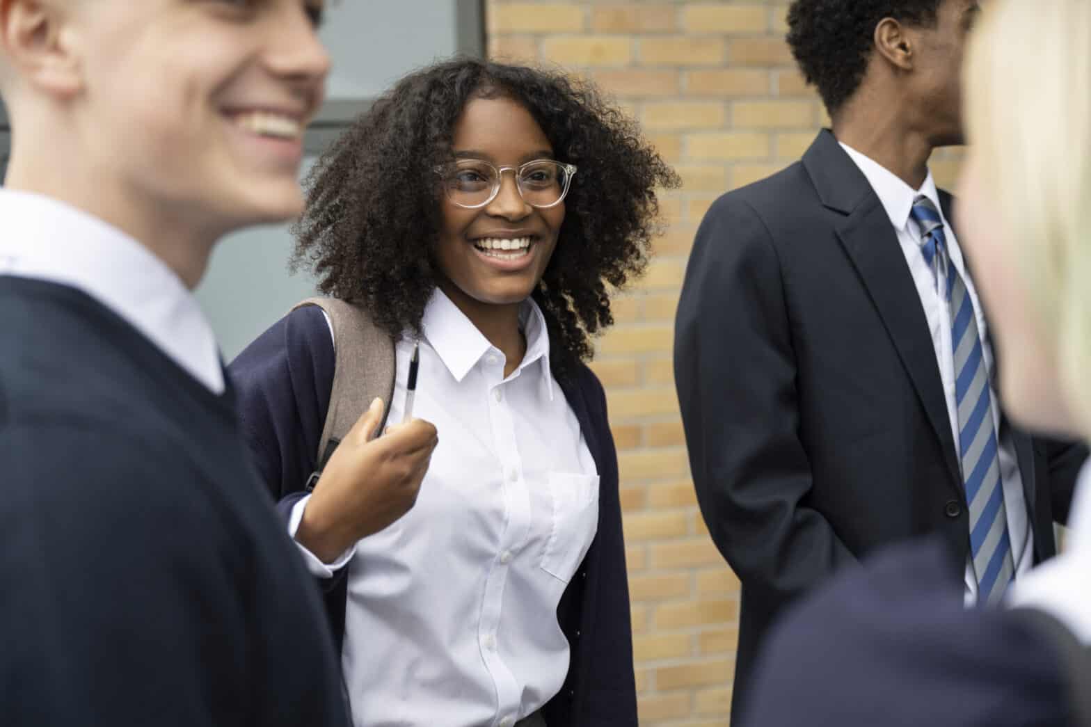 Candid view of cheerful teenage schoolgirl in uniform, eyeglasses, and backpack standing outdoors with friends, talking and laughing.