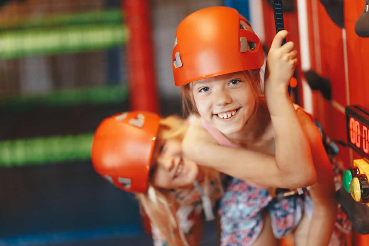 Two smiling young girls wearing hard hats on an indoor climbing wall.