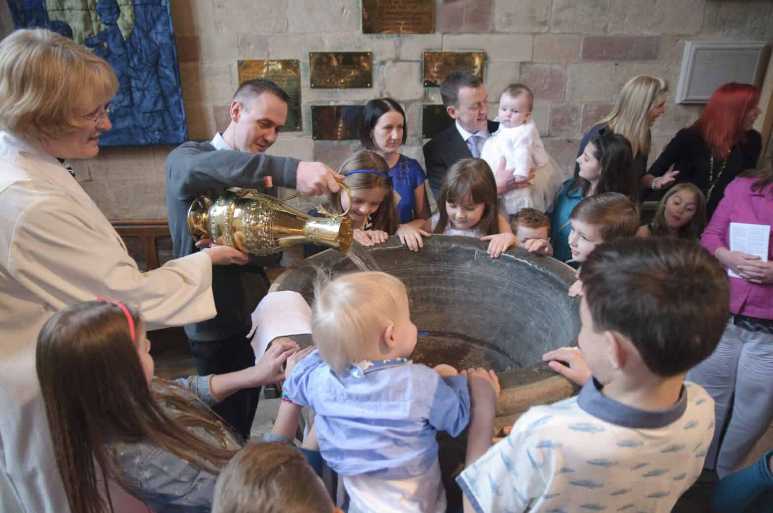 Water being poured into a font in church while children and adults crowd around.