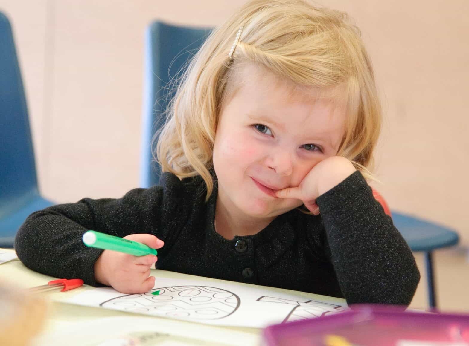 A young child holding a pen is sat at a table with a part finished colouring sheet. They smile at the camera.