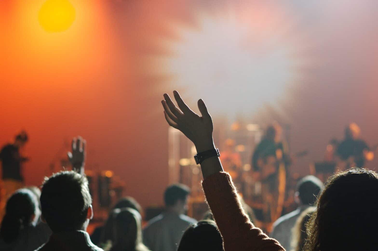 People with their hands up in praise during sung worship in a contemporary church. A worship band is playing on the stage in the background of the photo. Focus is on a woman's arm, hands outstretched.