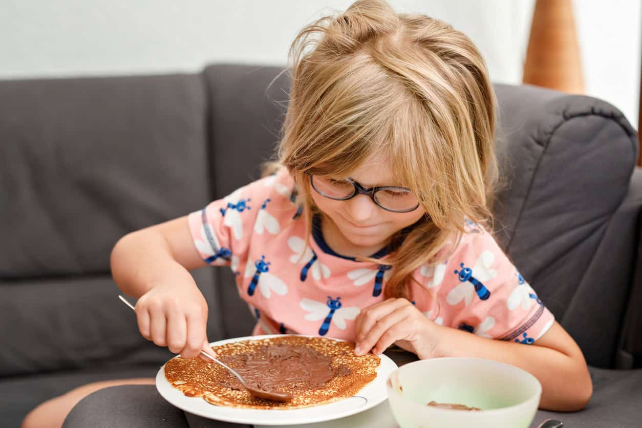 Young girl wearing glasses spreading chocolate on a pancake.