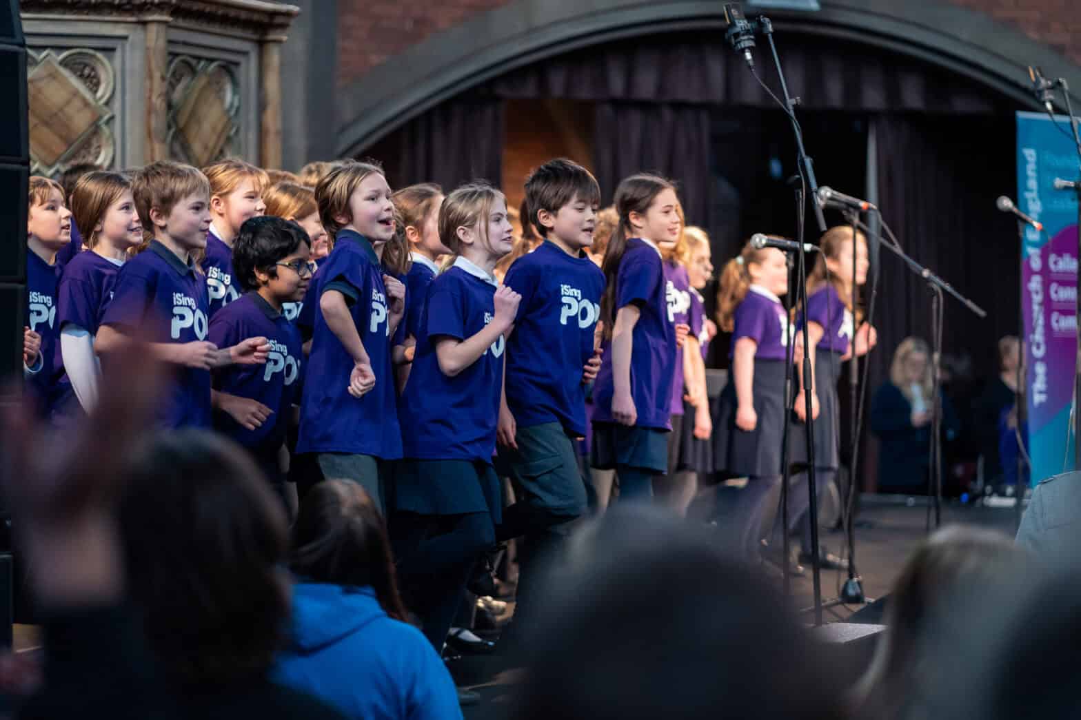 A group of school children on a stage in church singing, all wearing the same purple t-hirt with 'iSingPop' printed on the front.