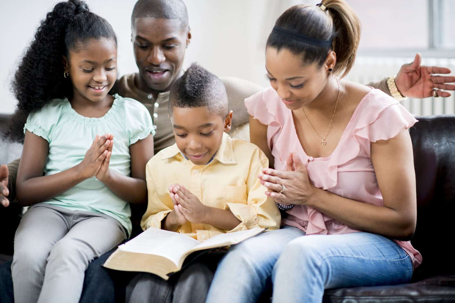 Happy family with mother, father and two children sitting on a sofa with a Bible and hands folded in prayer