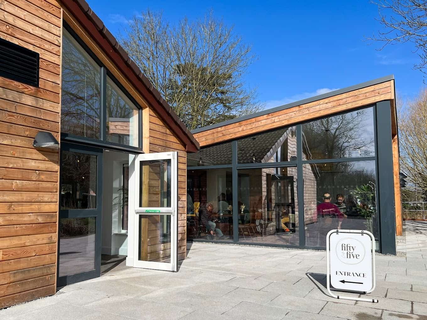 Wooden cafe building with large glass windows and an open door. The sun is shining though it seems to be winter because there are no leaves on the trees.