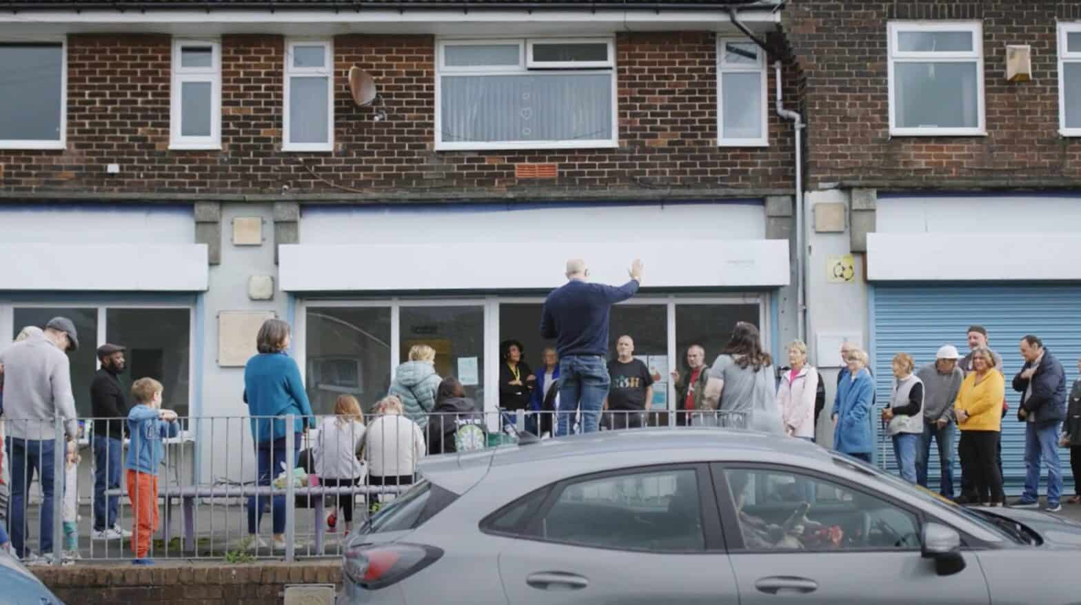 Lots of people standing outside a foodbank. There is a man standing on a bench addressing the people who are waiting.