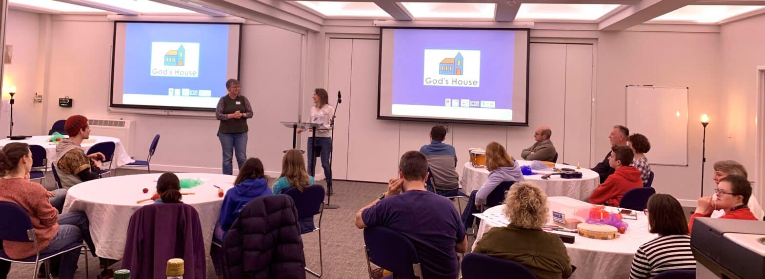 Four tables are set up in a room with seventeen people sitting around them. At the front of the room there is a microphone and two screens with slides for the worship. Everyone is paying attention and seems engaged.