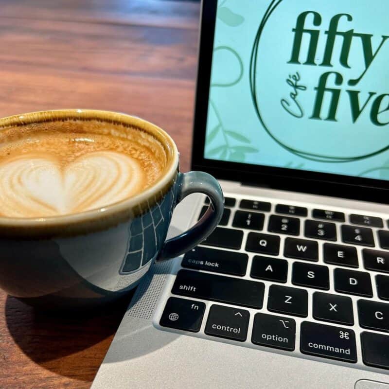 Stylish cup of coffee with a swirled heart on top and a laptop with Cafe Fifty-Five logo on the screen