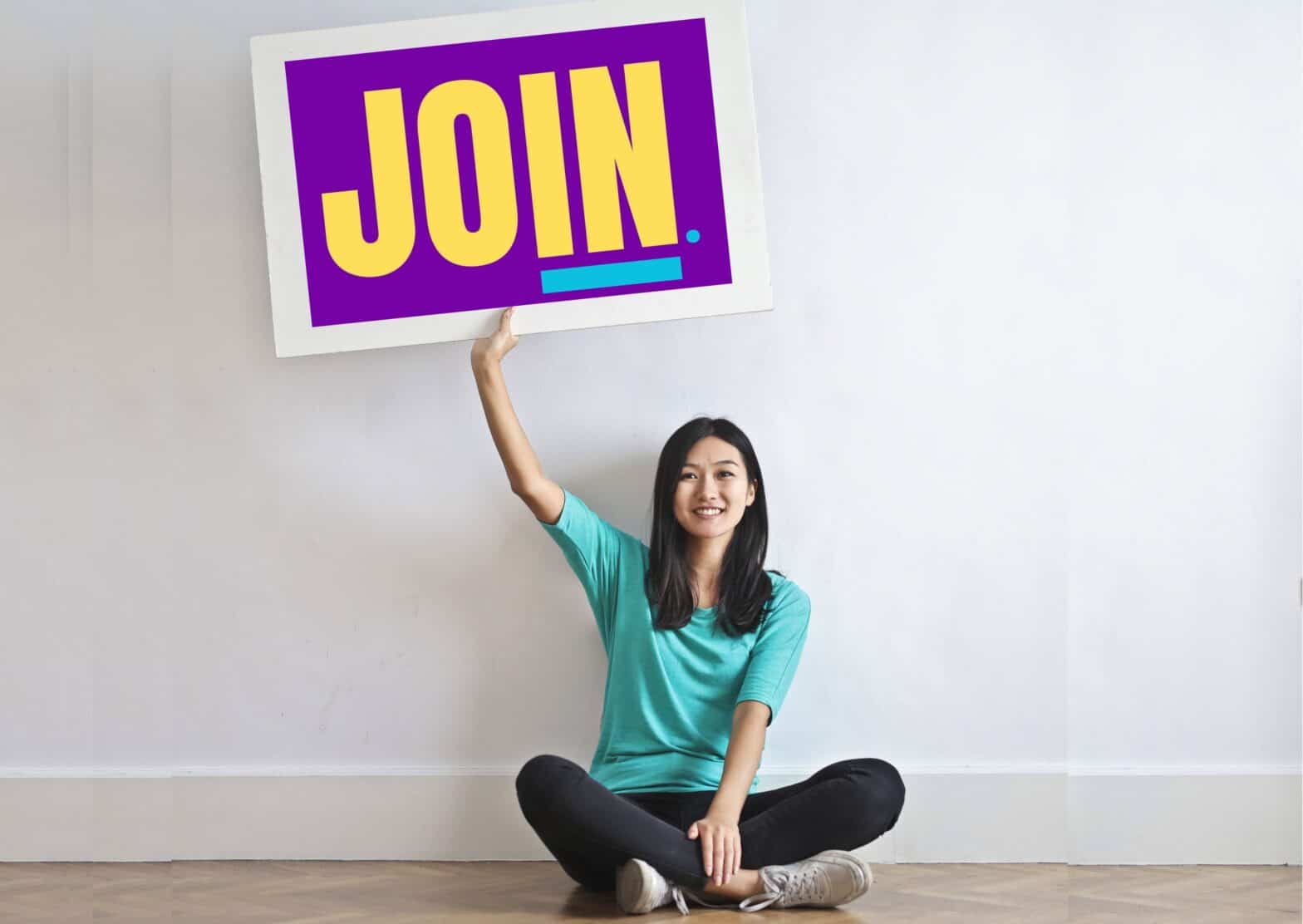 Girl sitting on floor holding a sign that says 'JOIN' in large yellow letters on a purple background