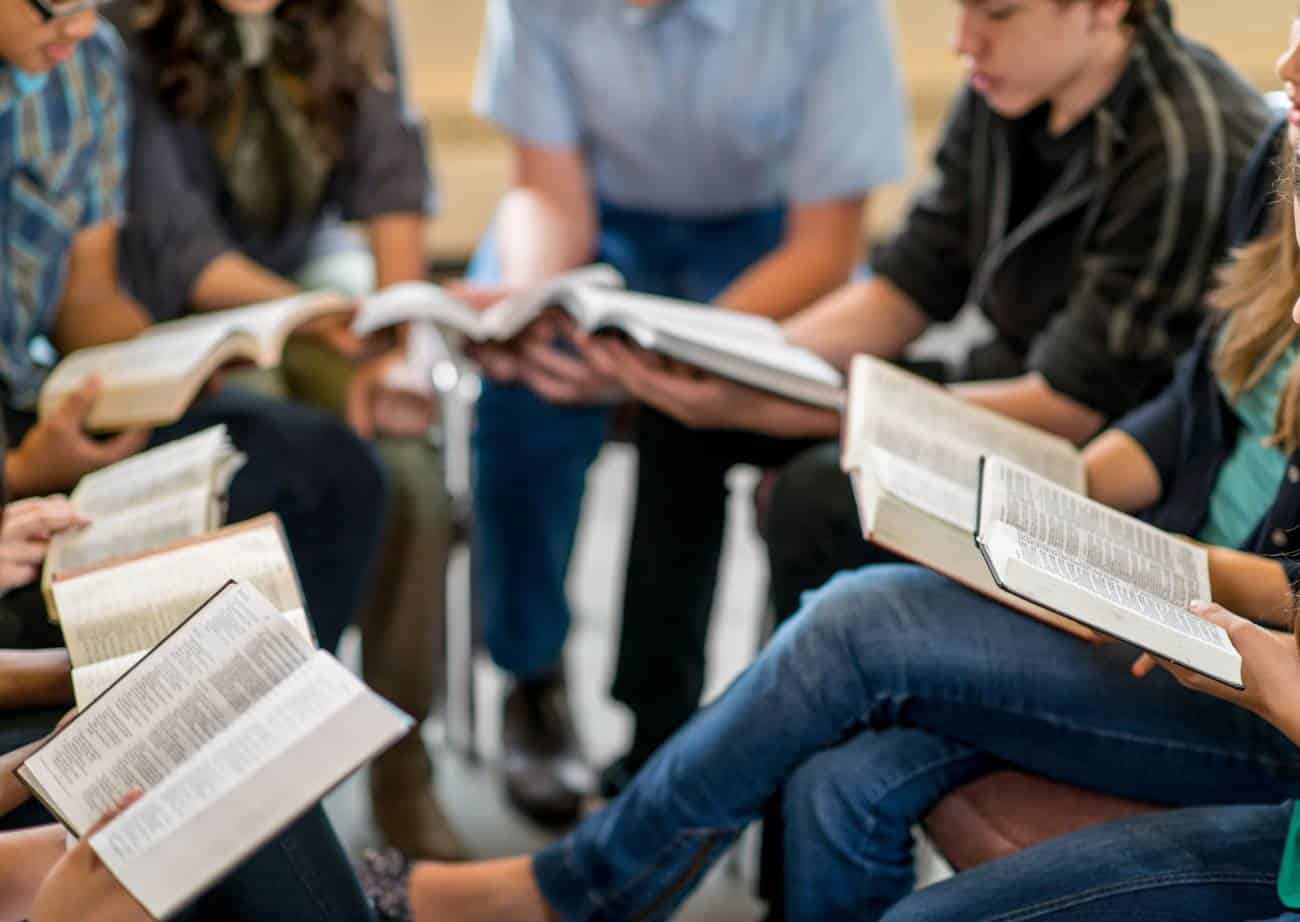 Group of people, possibly students, sitting around in a circle reading a book that may be the Bible.
