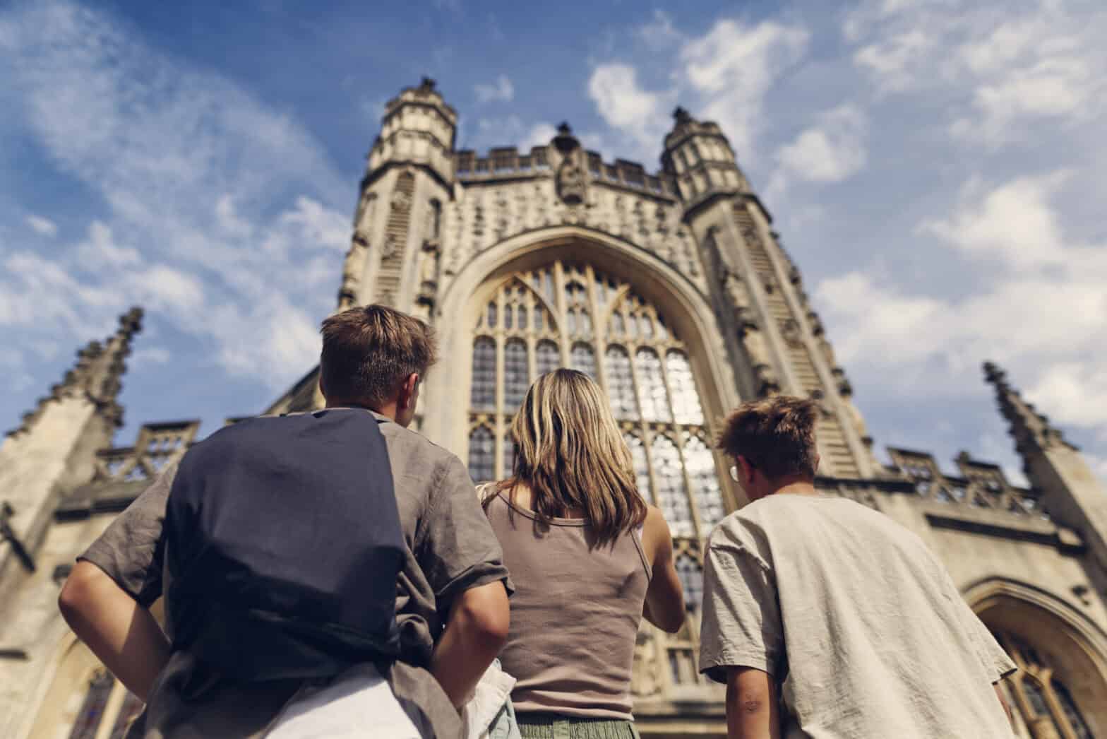 Three people standing outside of a church looking up in wonder. There is a blue sky dotted with clouds.