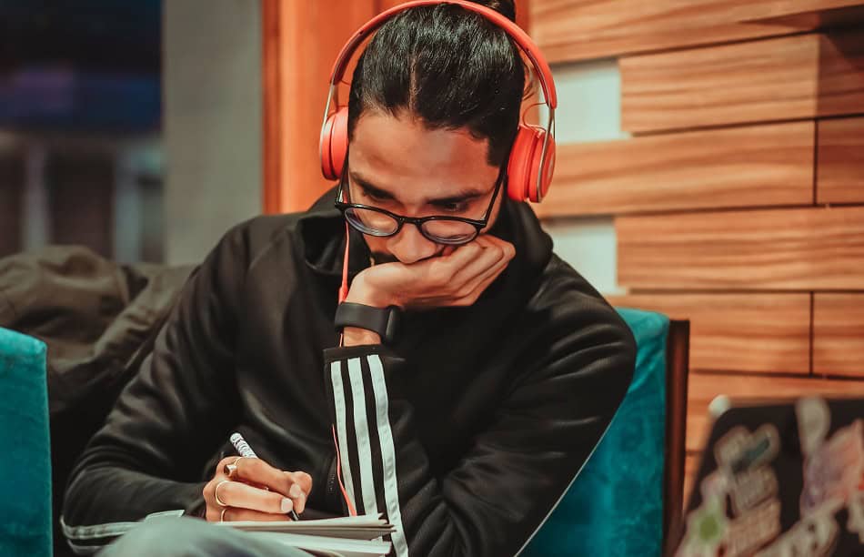 Man wearing orange headphones and writing notes whilst leaning his face on his hand.