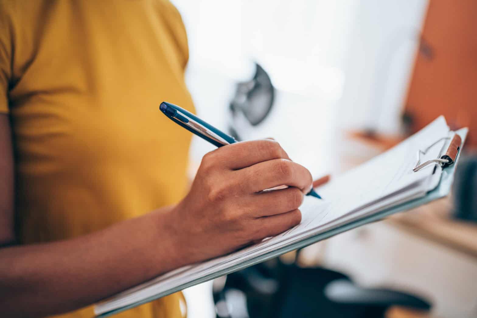 Woman in a yellow top making notes on papers secured on a clipboard.