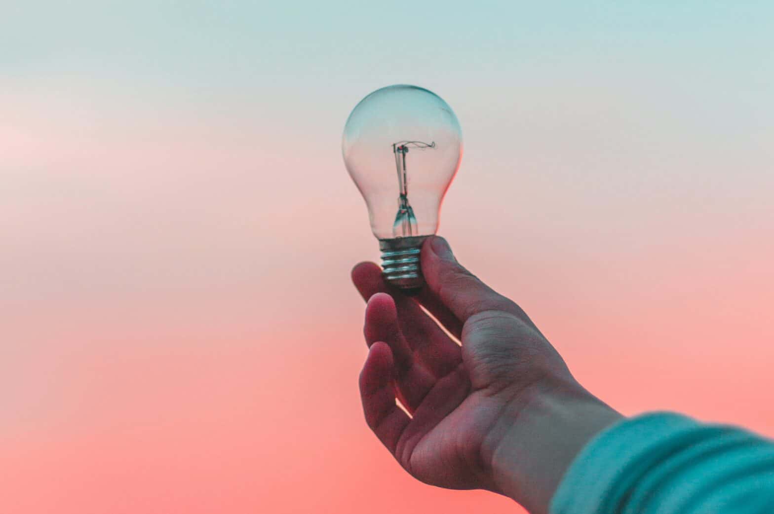 A hand holding a light bulb up to a sky that is half pink and blue