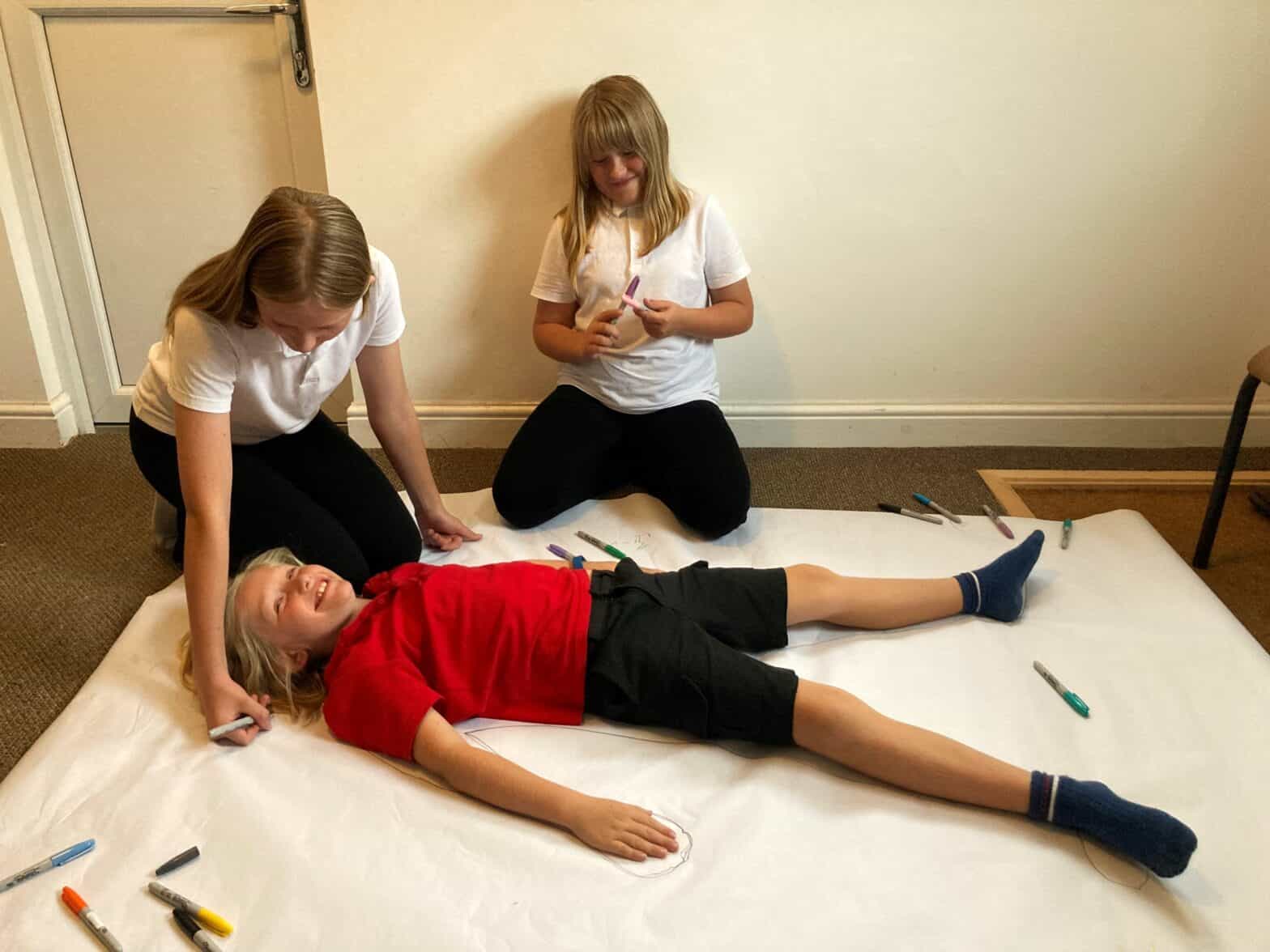 A child is lying on a piece of paper and being drawn around by two other children.