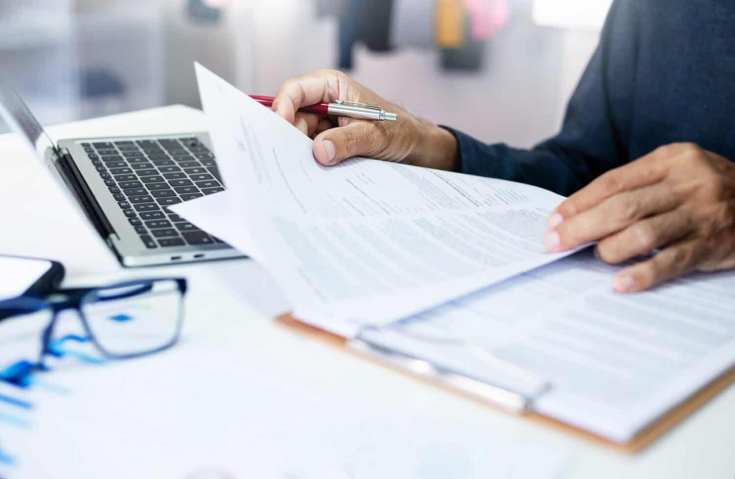 Image of a man's hands holding papers with a laptop and glasses on desk