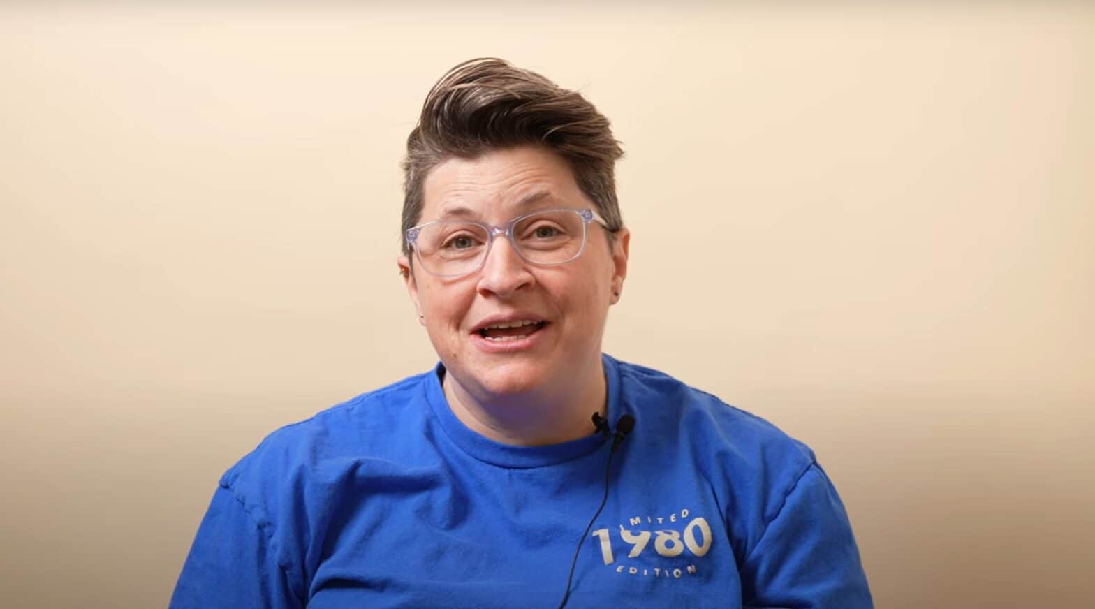 Person with glasses and blue t-shirt speaking against a peach background