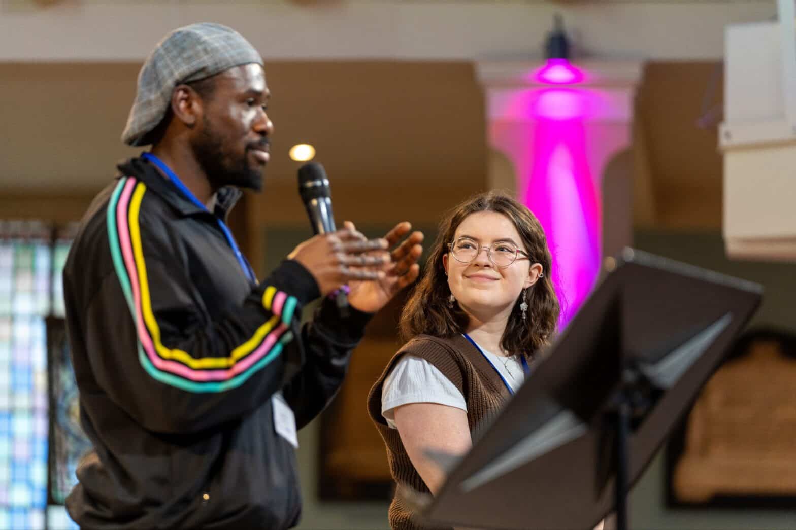 A man wearing a grey hat holiding a microphone in front of a lectern whilst a young woman looks at him smiling