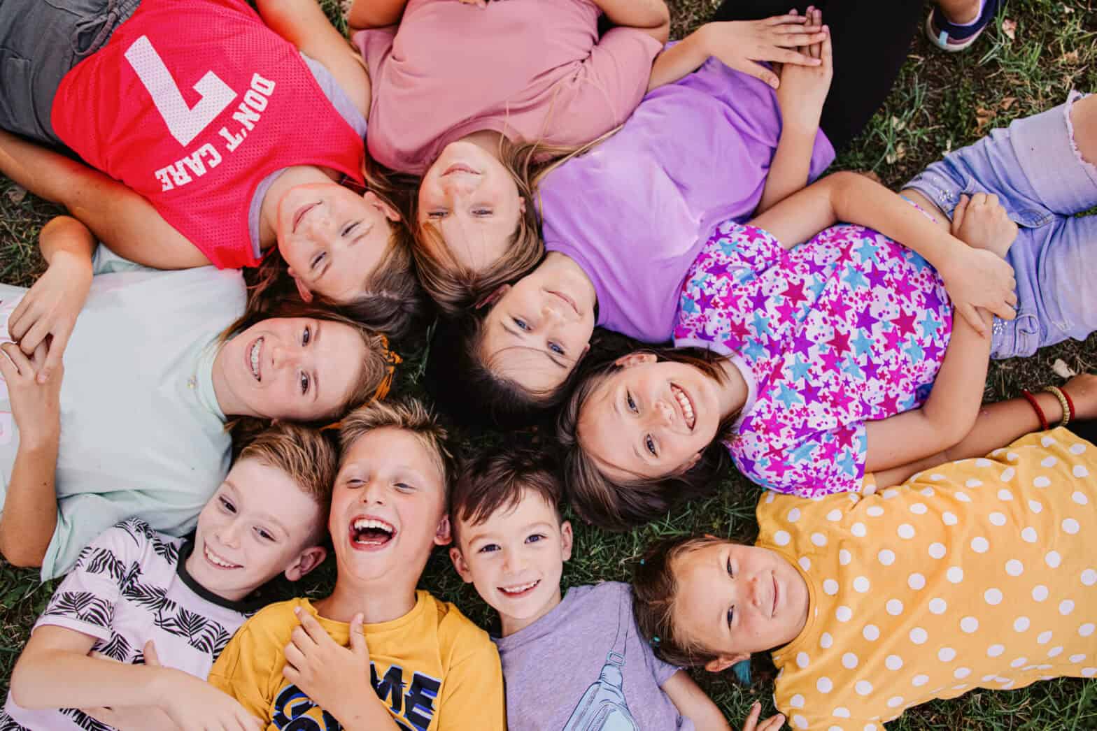 A group of young children, both girls and boys, laying on the grass looking upwards smiling to the camera