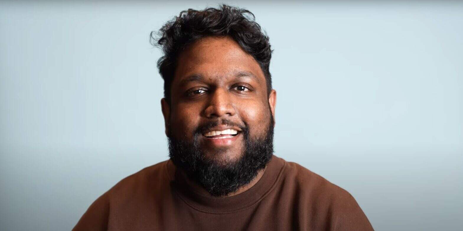 Man wearing brown top speaking against a blue background