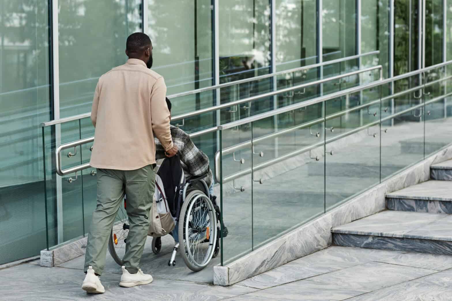 Someone pushing person in a wheelchair up a ramp next to a building with glass windows