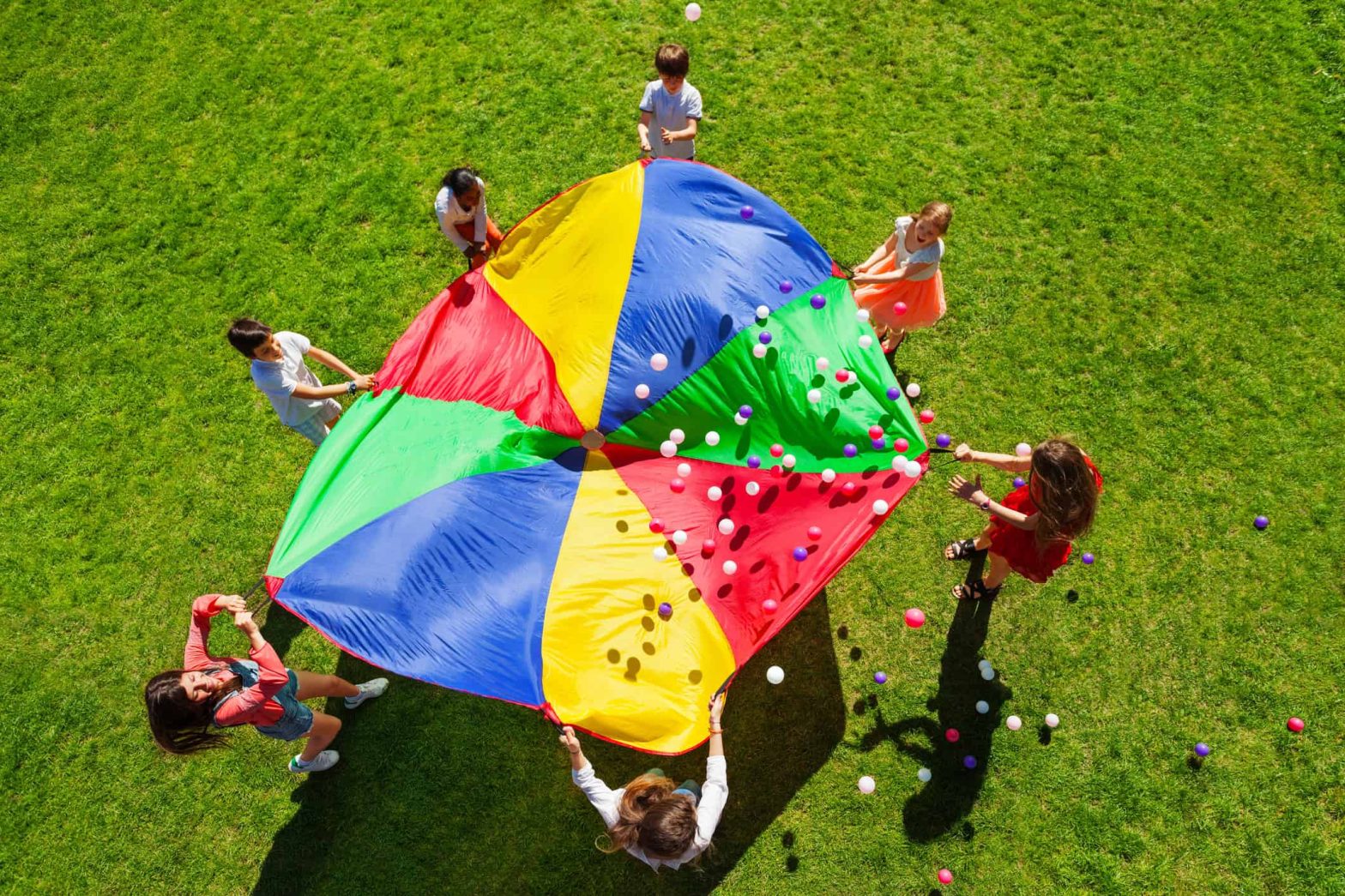 Top view picture of kids standing in a circle on a green lawn and holding a rainbow parachute full of colourful balls