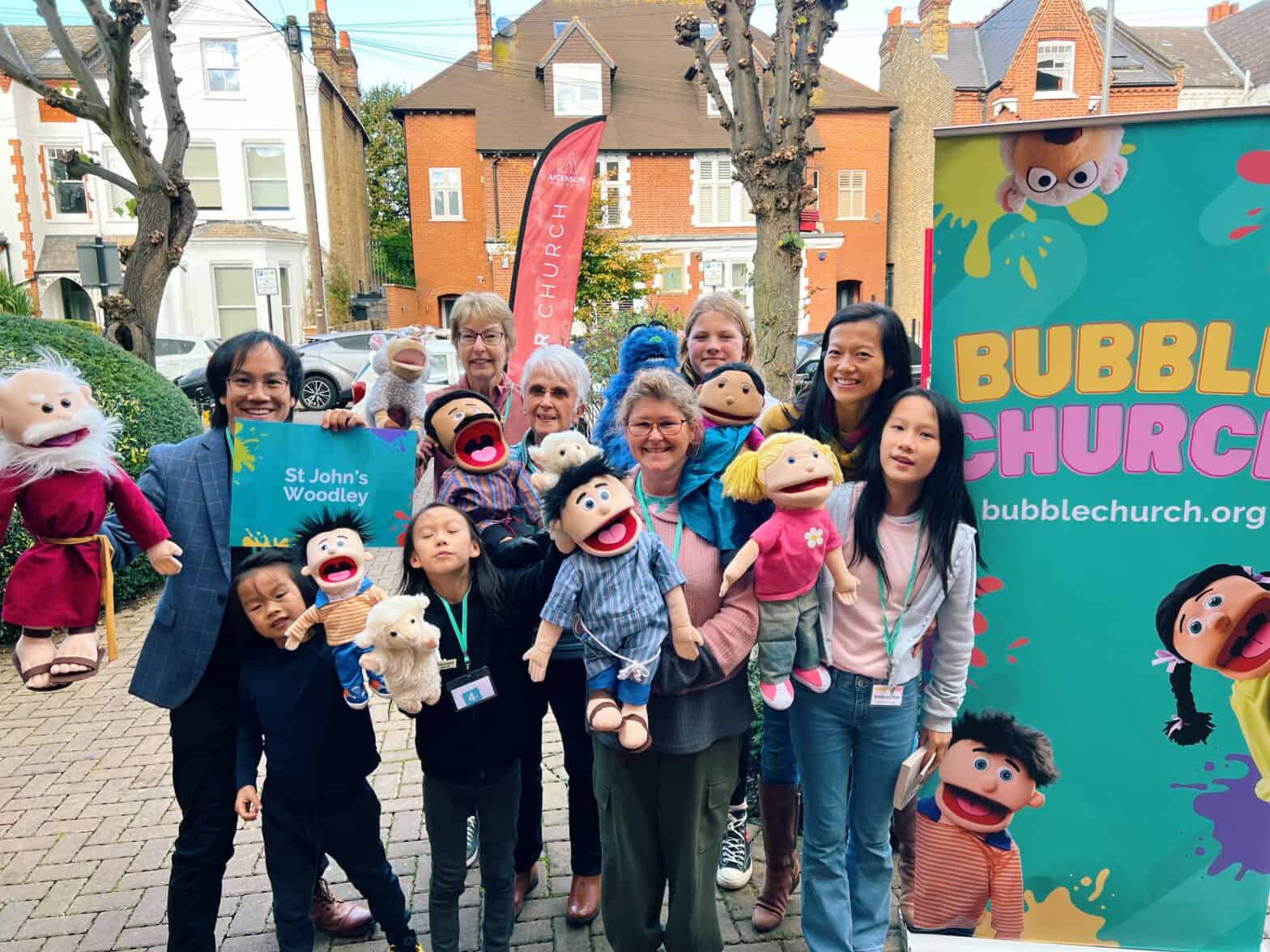 Group of adults and children holding puppet characters, posing in front of a "Bubble Church" banner. They are standing on a street in front of large residential buildings.
