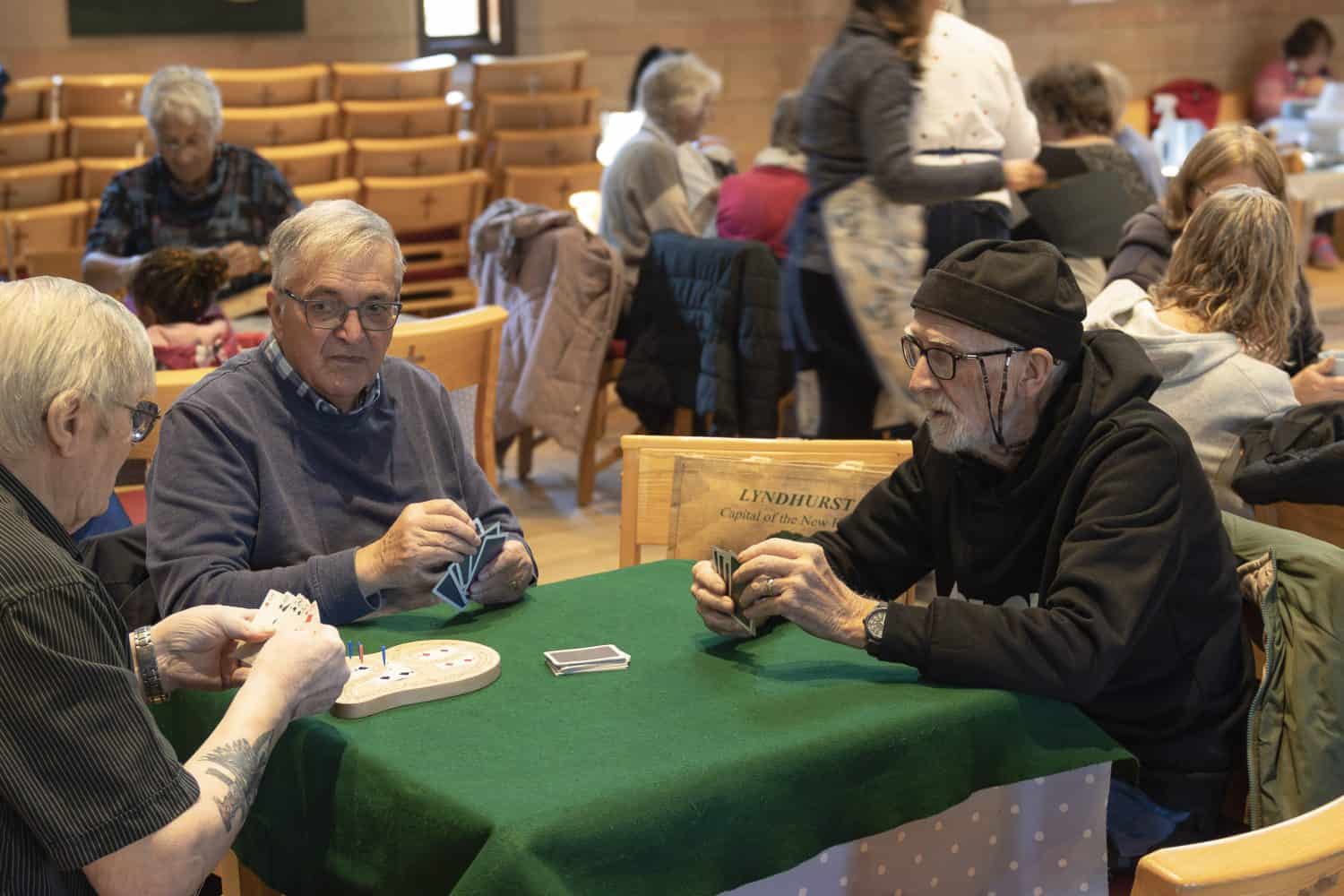 Three men playing cards and chatting at a table. There are lots of other people in the background having conversations and enjoying themselves at a Place of Welcome cafe.