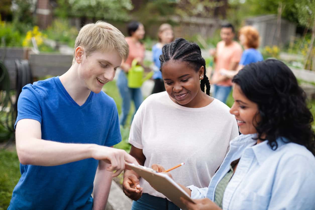 Three young adults standing outside are discussing something on a clipboard that they are all looking at