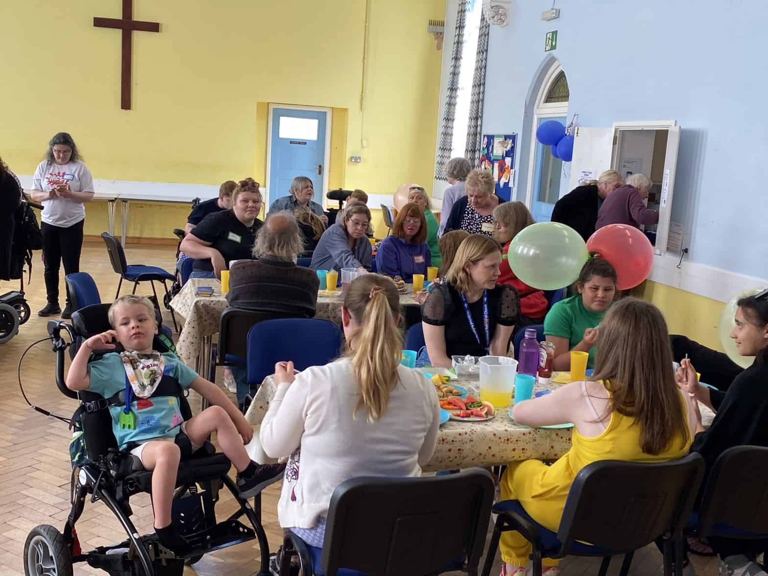 A church hall with yellow walls where several diverse people are sitting at tables eating and drinking. There are colourful balloons. A young boy sits in a disability pushchair.