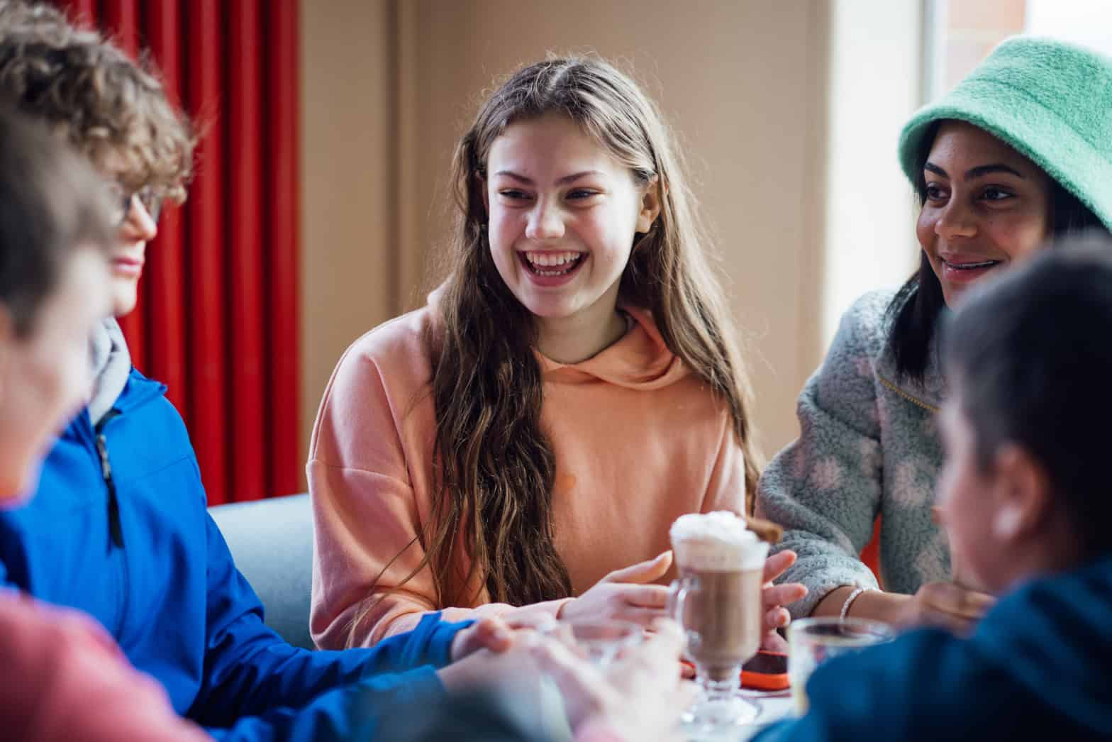 Five young people, male and female sitting indoors around a table drinking hot chocolate. A red curtain in background.