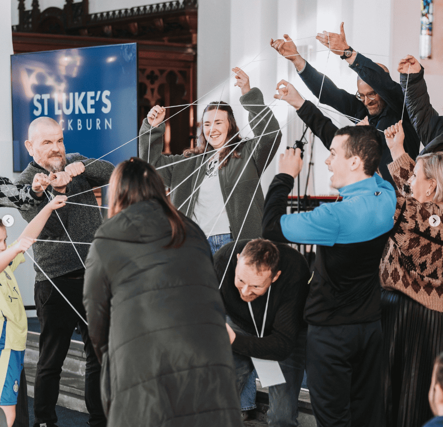 A group of adults are standing in a rough circle outside a white building. They are holding string around their hands forming a large cats cradle.