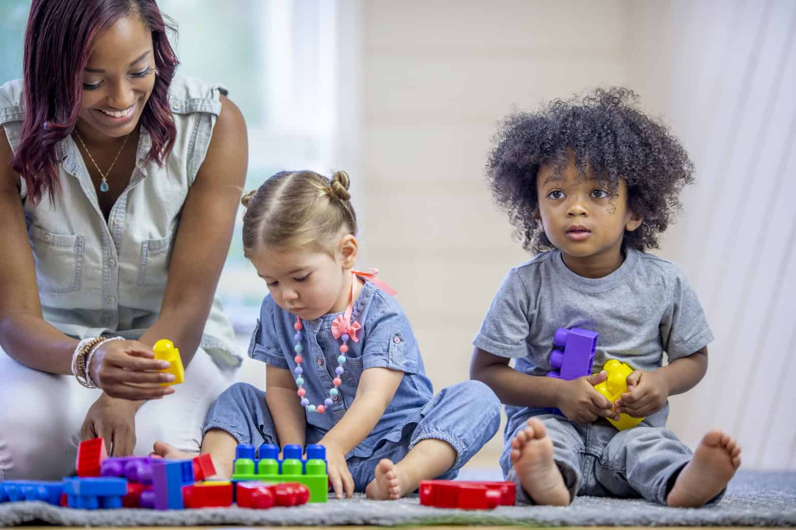 Two young children sitting side by side on the floor playing with brightly coloured building blocks. a lady accompanies them and is handing one of them a yellow block.