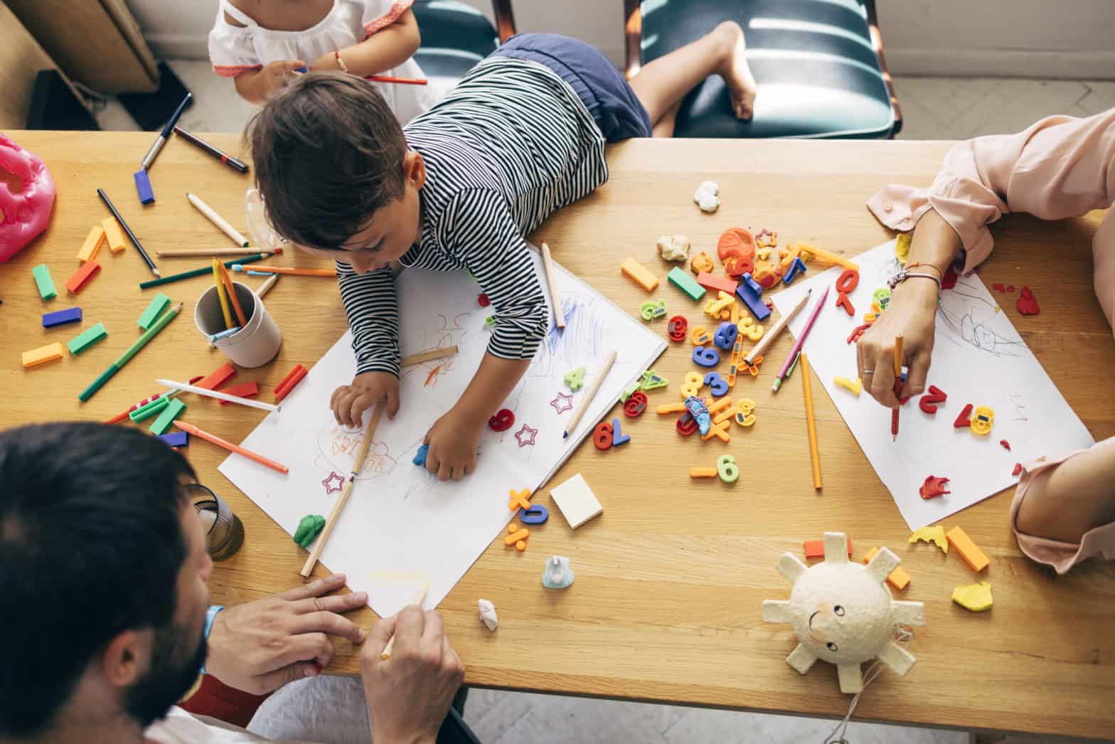 A young boy is on a table with lots of pens and magnetic letters . There is an adult helping him with craft.