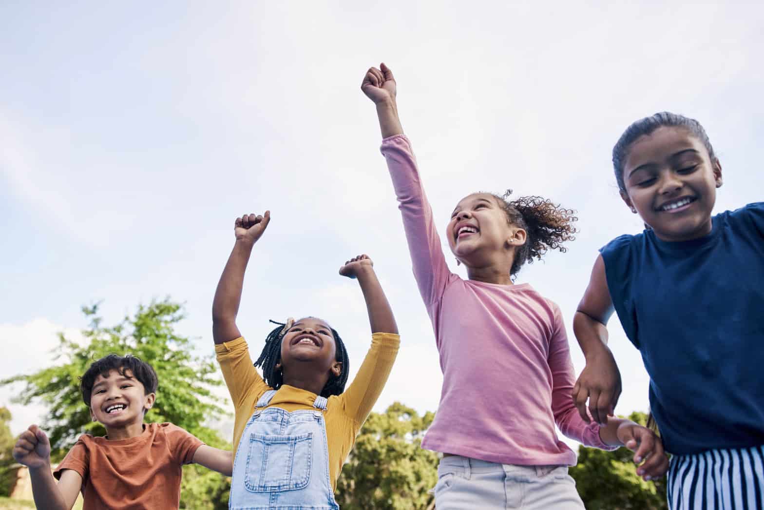 Four children, one is wearing a pink top and jeans. One is wearing a yellow top and demin dungarees. The other two are slightly out of shot. They are holding hands and jumping/smiling.