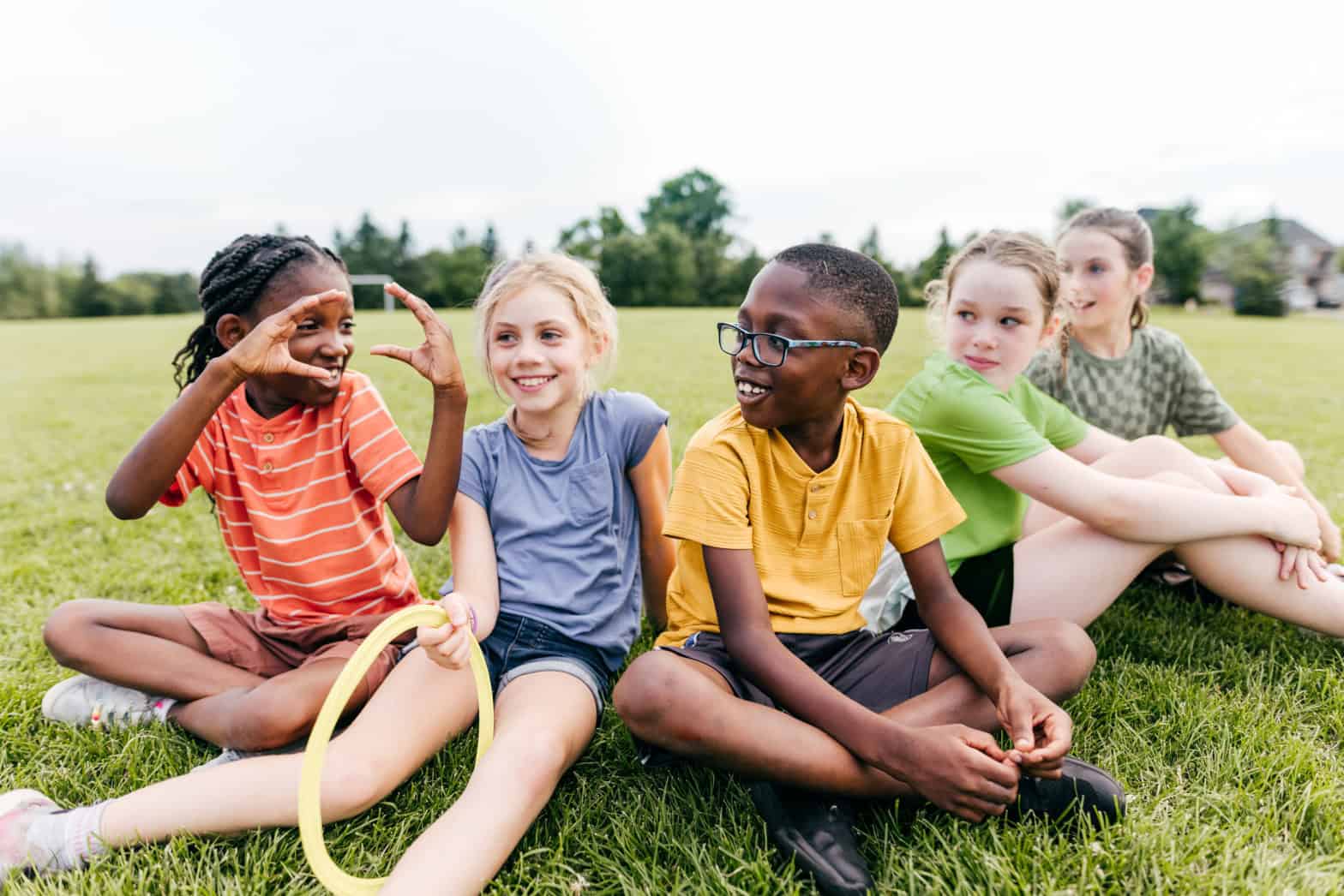 Five children sitting on grass wearing multi coloured tops. One child is holding her hands up forming a half circle whilst the others look at her smiling
