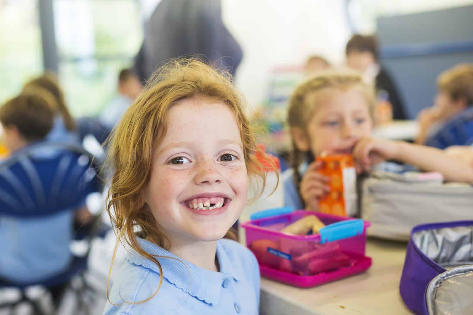 Young girl with red hair and a gap in her tooth sitting at a table smiling with her lunch and other peers in the background.